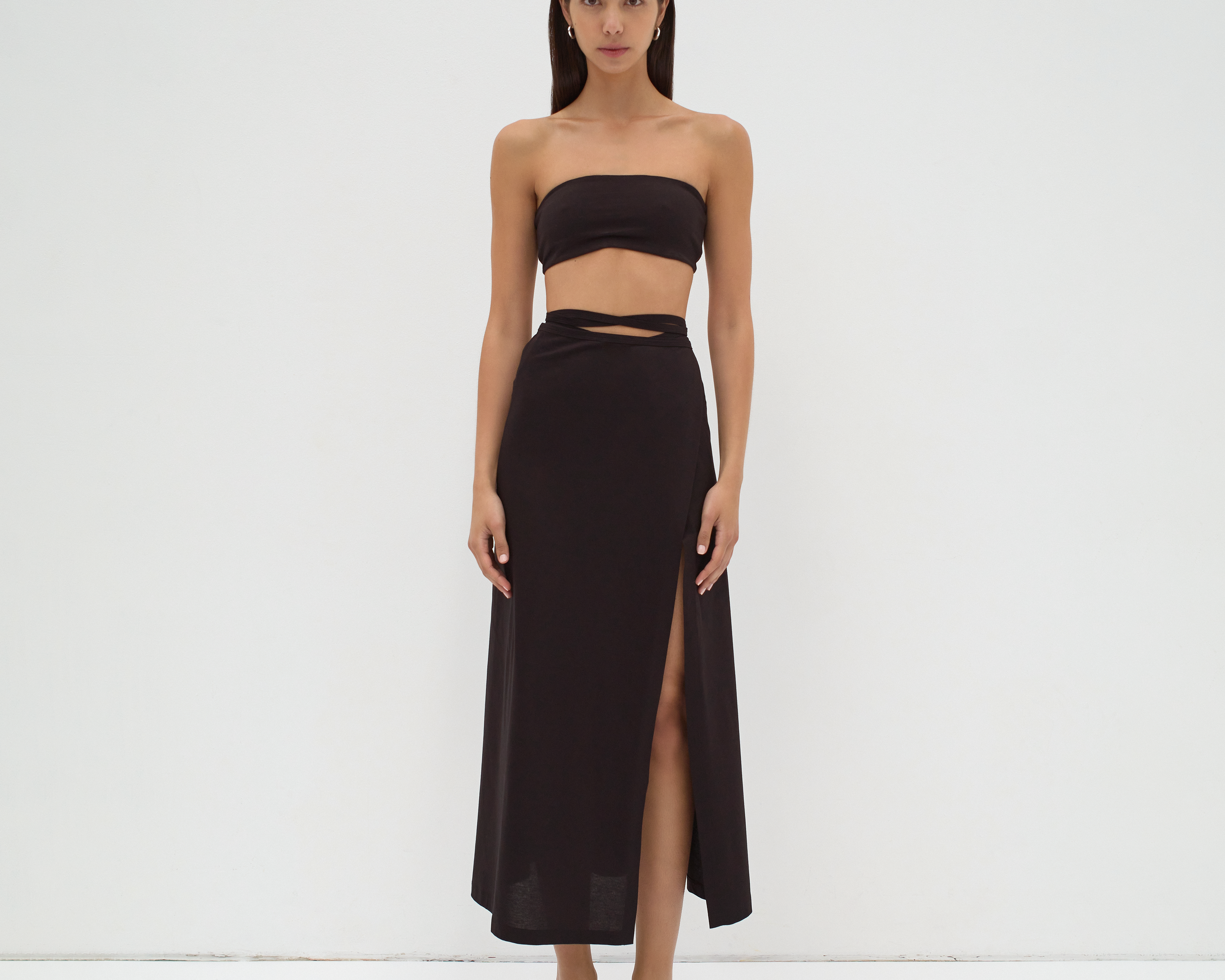 A model wearing a midi-length black skirt with extra-long ties and a split detail at the mid-thigh. The skirt is made from mercerised Turkish cotton by sener besim