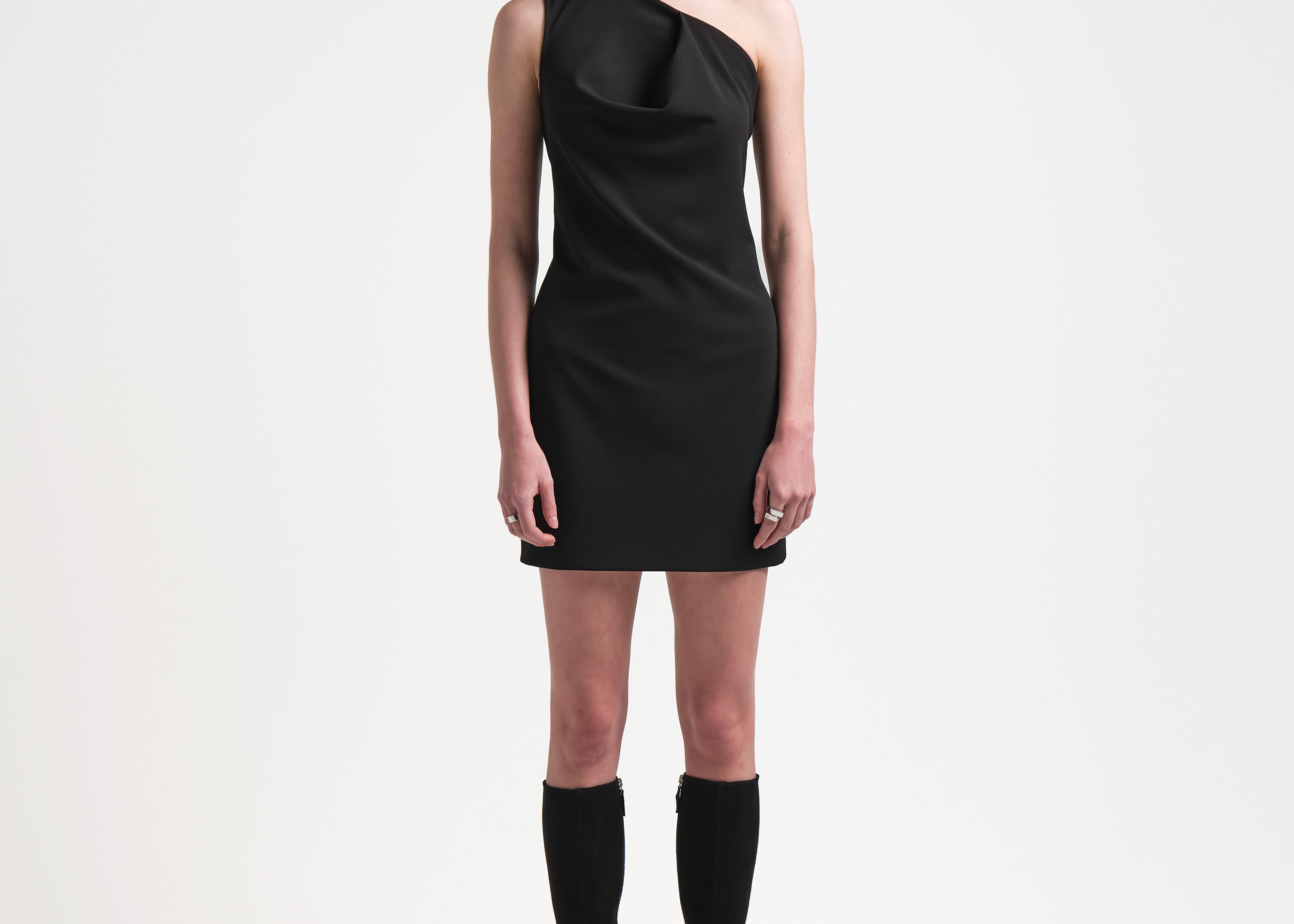 A model wearing a black one shouldered dress with an asymmetric neckline, designed to sit close to the body with a mini length. The dress is made from a mid-weight fabric and the model pairs it with long black boots.